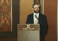 <span itemprop="name">John Crary speaking from a podium during an event...</span>